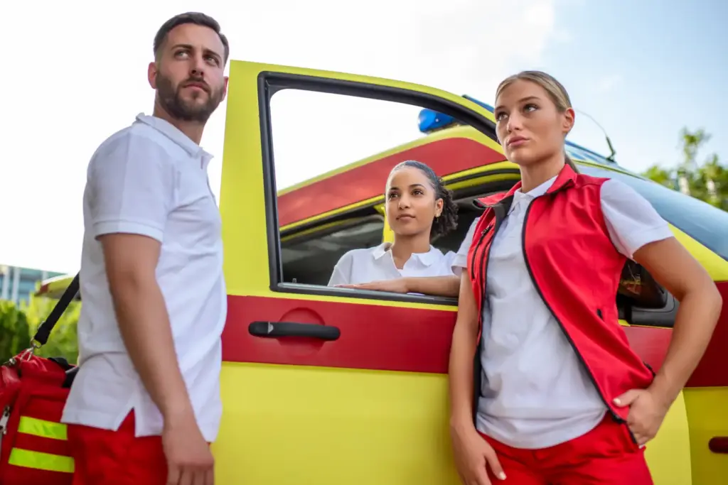 Ratownicy medyczni w czerwono-białych uniformach przy żółtej karetce, jeden z nich z torbą medyczną
