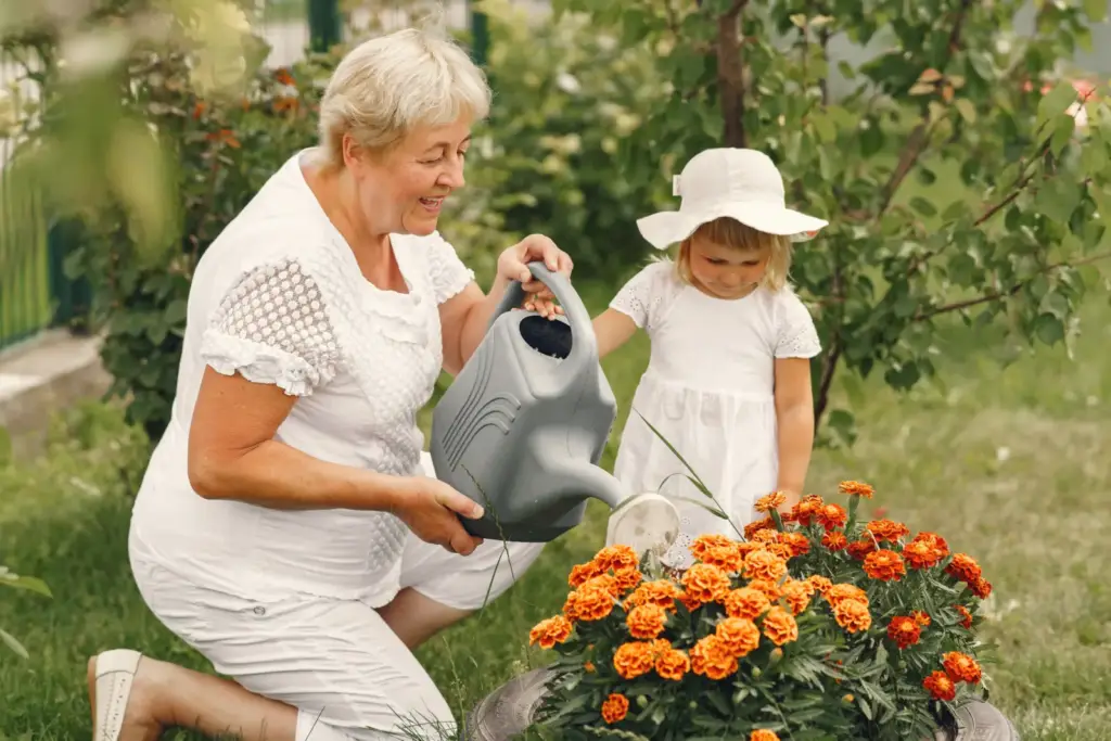 Babcia w białym kapeluszu podlewa kwiaty w letnim ogrodzie razem z małą dziewczynką