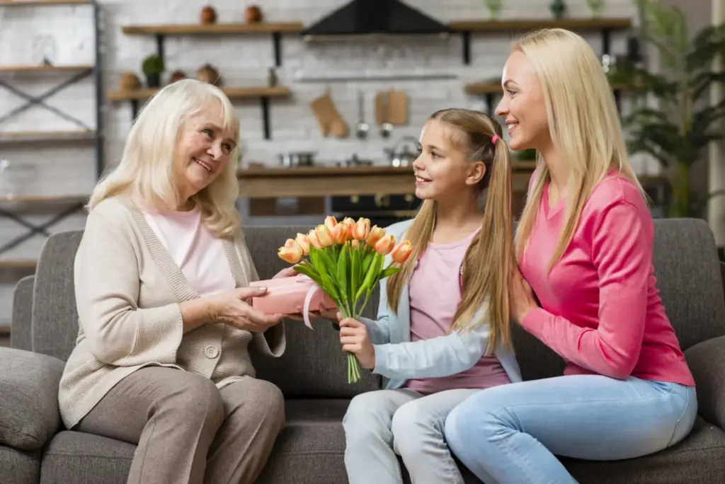 Dziewczynka wręcza starszej kobiecie bukiet tulipanów, obok siedzi uśmiechnięta mama